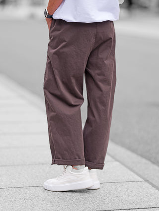Person trägt OFF#DLY Joggpant Melania schoko, locker geschnittene braune Hose mit umgeschlagenen Bündchen, kombiniert mit weißen Sneakers und grauen Socken. Smartwatch am linken Handgelenk sichtbar.