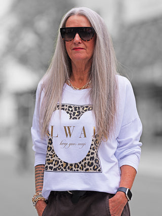 Frau trägt das OFF#DLY Longsleeve Always white mit goldenen Buchstaben und Leopardenmuster, kombiniert mit dunklen Hosen. Lässiger, moderner Look mit Statement-Print, perfekt für vielseitige Alltagsoutfits.