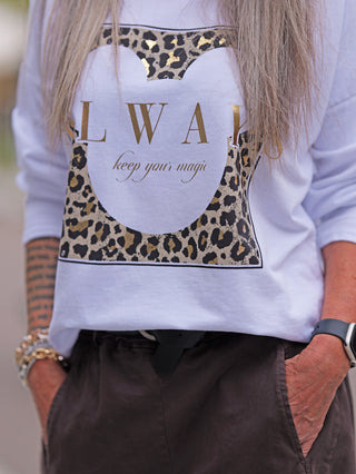 Nahaufnahme einer Person mit dem OFF#DLY Longsleeve Always white, featuring einem Leoparden-Mickey-Silhouette-Print und goldener Aufschrift LWAH keep your magic, kombiniert mit dunkler Hose und Accessoires.