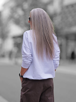 Person von hinten auf einer Stadtstraße, trägt das OFF#DLY Longsleeve Always white mit lockerem Schnitt und Statement-Frontprint. Kombiniert mit dunkler Hose und Smartwatch, vermittelt es lässigen Chic.