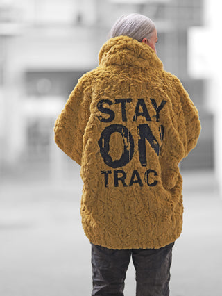 Person von hinten in urbaner Umgebung, trägt #VDR Mantel 18185 pistacio, eine flauschige, oversized Kuscheljacke mit auffälligem Rückendruck STAY ON TRAC in Schwarz.