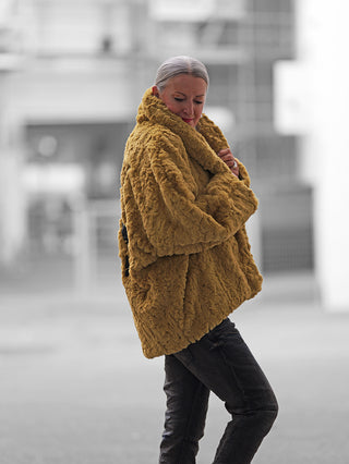 Frau in urbaner Umgebung trägt #VDR Mantel 18185 pistacio, einen oversized, flauschigen Teddy-Mantel mit hohem Kragen, während sie ihn anpasst. Ihr silbernes Haar ist zurückgebunden.