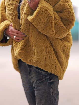 Person trägt #VDR Mantel 18185 pistacio, eine oversized Kunstpelzjacke mit hohem Kragen. Hände sind mit Ringen verziert, eine hält den Mantel, die andere ruht an der Taille.