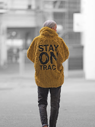 Person in urbaner Kulisse trägt #VDR Mantel 18185 pistacio, ein übergroßer, kuscheliger Faux-Fur-Mantel mit auffälligem Rückenprint STAY ON TRAC, perfekt für winterlichen Streetstyle.