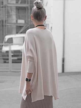 Frau von hinten in urbaner Umgebung, trägt den OFF#DLY Pulli Olivia Creme: oversize, creamfarben, seitliche Schlitze, über einem langärmeligen Top, kombiniert mit einem neutralen Faltenrock und Accessoires.