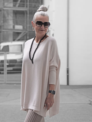 Ältere Frau trägt den OFF#DLY Pulli Olivia Creme, ein locker geschnittenes, cremefarbenes Oberteil mit gerippten Ärmeln und seitlichen Schlitzen, kombiniert mit stilvollen Accessoires für einen eleganten Casual-Look.