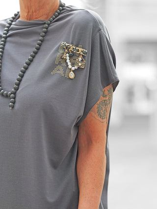 BSB Pulli Carbon: Lockeres, schiefergraues Kurzarmshirt mit einer seitlichen Brosche aus Perlen und Anhängern. Das Design betont die feine Materialqualität und den einzigartigen Stil des Shirts.