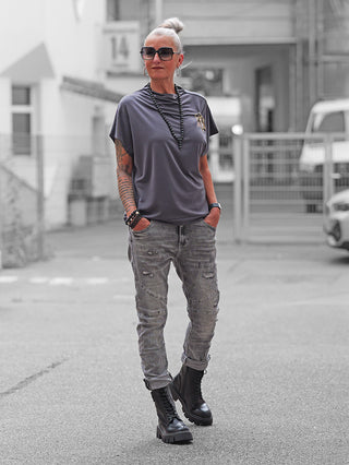 Ältere Frau im BSB Pulli Carbon, einem locker sitzenden grauen Kurzarmshirt, kombiniert mit distressed Jeans, schwarzen Perlen, Tattoos und klobigen Stiefeln.