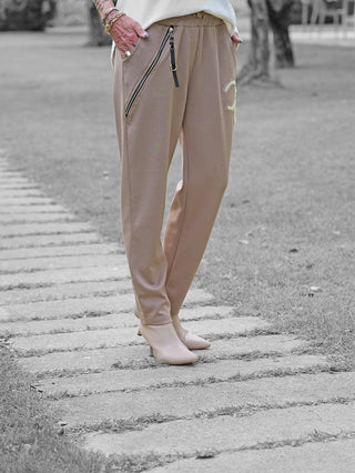 Person trägt OFF#DLY Joggpant Bonny beige mit diagonalem Reißverschlussdetail und Perlenlogo. Die Hose kombiniert Komfort und Glamour, ergänzt durch passende Stiefeletten und Schmuck an den Händen.