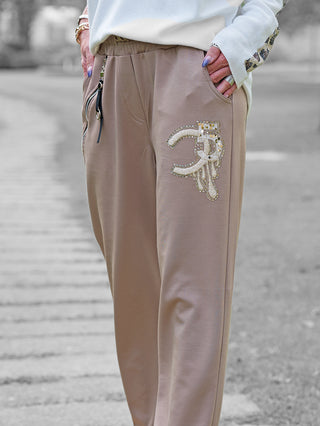 Nahaufnahme der OFF#DLY Joggpant Bonny beige mit elastischem Bund, seitlichen Taschen, funkelnder Strass-Applikation 35 und Reißverschluss-Detail, getragen mit weißem Oberteil und Accessoires.