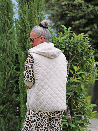 Person im Garten trägt die OFF#DLY Steppweste Bella beige, eine leichte, gesteppte Kapuzenweste über einem Leoparden-Top. Silbernes Haar im Dutt, Sonnenbrille; sportlich-eleganter Look.
