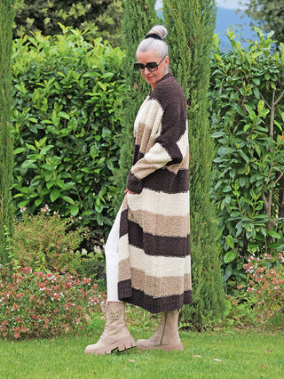 Frau trägt den OFF#DLY Cardigan Sandy brown Stripes, einen langen, gestreiften Cardigan in warmen Naturtönen mit offenen Schnitt und seitlichen Taschen, kombiniert mit weißen Hosen und beigen Stiefeletten.