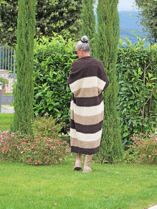 Person von hinten im Garten, trägt den OFF#DLY Cardigan Sandy brown Stripes mit langen, dicken Streifen und offenem Schnitt. Die Strickjacke hat einen gemütlichen Look, ideal für kühle Tage.