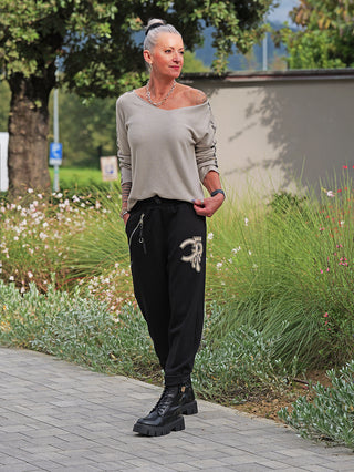 Frau trägt OFF#DLY Joggpant Bonny black, schwarze Jogger mit Strassapplikation am Oberschenkel, kombiniert mit grauem Pullover und schwarzen Stiefeln, auf einem Weg mit Pflanzen im Hintergrund.