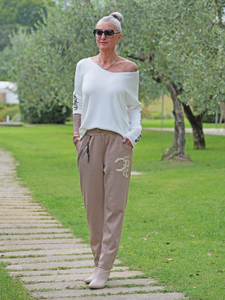 Eine Frau trägt die OFF#DLY Joggpant Bonny beige mit funkelnder Strass-Applikation und Reißverschlusstaschen, kombiniert mit einem weißen Pullover und beigen Stiefeletten auf einem Steinweg.