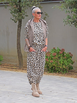 Ältere Person im Freien trägt die OFF#DLY Steppweste Bella taupe, leicht glänzend, mit Kapuze und Druckknöpfen, über einem Leoparden-Outfit, kombiniert mit Kampfstiefeln und oversized Sonnenbrille.