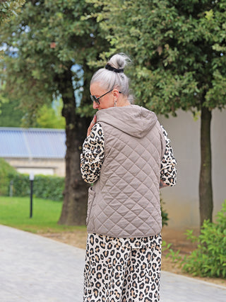 Person von hinten in OFF#DLY Steppweste Bella taupe, eine gesteppte Kapuzenweste mit Druckknöpfen und Taschen, über einem langärmeligen Oberteil und Leopardenmuster-Hose im Garten stehend.
