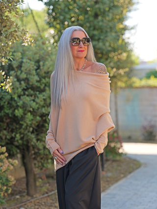 Frau in OFF#DLY Pulli Irene asymetric camel, locker geschnitten, schulterfrei, zeigt Tattoo, kombiniert mit schwarzer Hose, Statement-Kette und Lavendelnägeln, steht auf einem Weg mit grüner Umgebung.