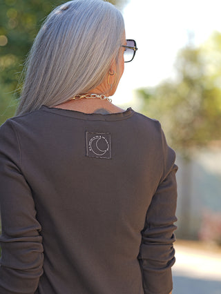 Person von hinten mit langen, silbernen Haaren, trägt das OFF#DLY LS Alice schoko mit schmalem Schnitt und Label-Patch. Goldkette und Ohrringe ergänzen den Look.