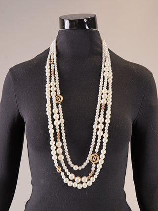 Die OFF#DLY Kunstperlenkette 3 Strang white/gold zeigt lange, gestufte weiße Perlen mit goldfarbenen Verbindungen und runden Anhängern auf einem schwarzen Kleiderbüsten-Torso, ideal als Statement-Schmuck.