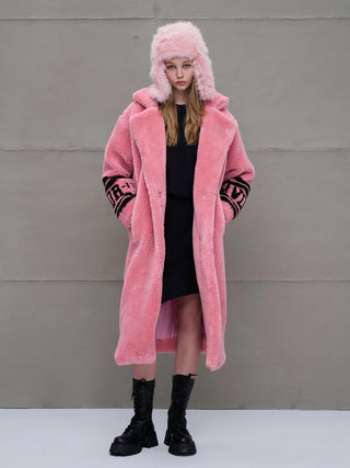 Junge Frau trägt #VDR Coat pink, einen langen, plüschigen, rosa Faux-Fur-Mantel mit breitem Kragen, kombiniert mit passendem Trapper-Hut und klobigen schwarzen Stiefeln, stehend vor grauer Wand.