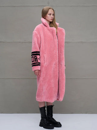Eine junge Frau trägt den #VDR Coat pink, einen langen, plüschigen Faux-Shearling-Mantel mit hohem Kragen und schwarzen Grafikdetails am Ärmel, kombiniert mit klobigen schwarzen Stiefeln.