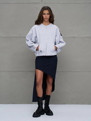 Frau trägt #VDR Sweatjacke grey mel mit Reißverschluss, weiten Ärmeln und elastischem Saum. Die Hände in den Taschen, kombiniert mit asymmetrischem Rock und robusten Stiefeln, urbaner Streetstyle-Look.