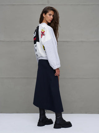 Junge Frau trägt #VDR Sweatjacke grey mel, kombiniert mit dunkelblauem Rock und schwarzen Boots; Jacke mit markantem Streetstyle und Fallschirmstoff-Details auf der Rückseite, moderne urbane Silhouette.