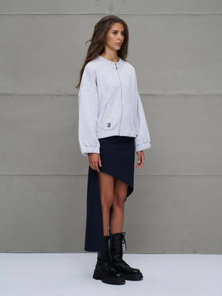 Junge Frau in Profil, trägt #VDR Sweatjacke grey mel mit Reißverschluss, Balloon-Sleeves und Taschen, kombiniert mit asymmetrischem Rock und Stiefeln, präsentiert urbanen Streetstyle vor grauer Wand.