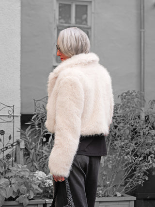 Eine Person trägt die OFF#DLY Felljacke Luisa white, ein kurz geschnittenes, cremefarbenes Kunstpelz-Jackett, kombiniert mit einer schwarzen Tasche, ideal für moderne Herbst- und Winterlooks.