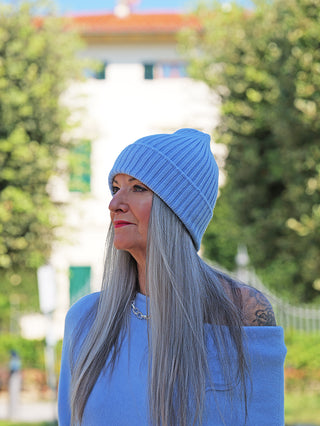 Frau im Profil trägt die OFF#DLY Mütze Anna hellblau, eine gerippte, hellblaue Strickmütze. Ihr langes, graues Haar fällt über einen passenden, schulterfreien Pullover.
