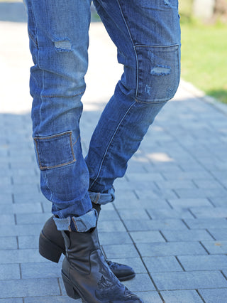 Nahaufnahme von unteren Beinen in hochgekrempelten, blau zerstörten OFF#DLY Denim Ines Midblue Destroy Jeans mit Patchdetails, kombiniert mit schwarzen Lederstiefeletten auf einem gepflasterten Gehweg.