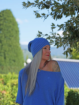 Frau mit langen grauen Haaren trägt die OFF#DLY Mütze Anna denimblue, eine gerippte Strickmütze, kombiniert mit einem schulterfreien Top, vor einer Gartenkulisse unter einem Blattzweig.