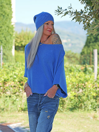 Person trägt die OFF#DLY Mütze Anna denimblue im Freien, mit lässigem Look, kombiniert mit einem schulterfreien blauen Pullover und Jeans, vor einer grünen Kulisse.