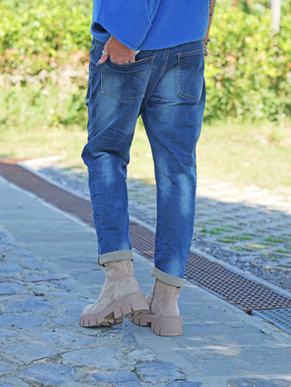 Person mit umgeschlagenen blauen OFF#DLY Denim Dunja destroy Midblue Jeans, lässiger Schnitt und dezente Destroyed-Details, trägt helle Wildlederstiefeletten auf Steinweg, rechte Hand in der Tasche.