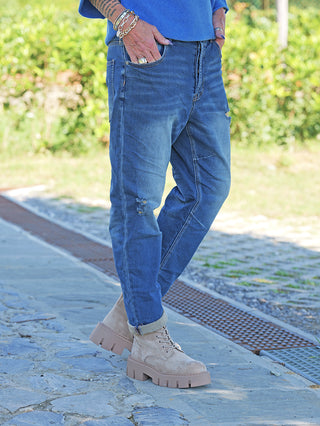 OFF#DLY Denim Dunja destroy Midblue: Cuffed, leicht zerstörte blaue Jeans mit entspanntem Schnitt und moderner Waschung, getragen mit beigefarbenen, klobigen Stiefeletten auf einem Steinweg.