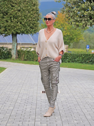 Frau trägt OFF#DLY Cardigan Safira stone, lockerer Strick mit tiefem V-Ausschnitt und großem Knopf, kombiniert mit Metallic-Taupe-Cargohose und beigen Stiefeletten, elegant-lässiger Look.