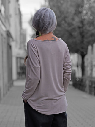 Person von hinten in OFF#DLY Bea taupe Longsleeve mit lockerer Passform, tiefem V-Ausschnitt, sichtbarer Rückentätowierung. Kombiniert mit dunkler Hose, auf urbaner Straße spazierend.