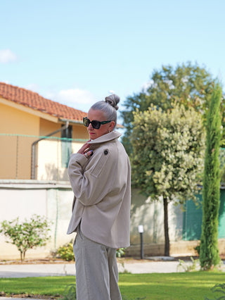 Frau trägt die OFF#DLY Jacke Valeska creme mit klassischem Reverskragen und doppelter Knopfleiste, kombiniert mit lockeren hellen Hosen, stehend in einem Garten.