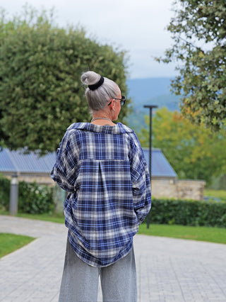 Ältere Person trägt die OFF#DLY Bluse Greta blau kariert, zeigt Rückansicht mit silbernem Haarknoten, Sonnenbrille, Kettenhalskette, kombiniert mit hellgrauen Hosen auf einem gepflasterten Parkweg.