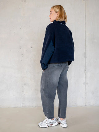 Frau trägt Penn&Ink Denim WZ25811 grey Barrel-Jeans mit elastischem Bund und verstellbarem Saum in einem schlichten Studio, kombiniert mit einem navy-blauen Fleece-Jacke und weißen Sneakers.
