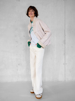 Frau in Penn&Ink Jacke W25c321 ecru, ein shearlinggefütterter Bomber, trägt lässig Hände in Taschen, kombiniert mit grauem Shirt, off-white Hose und passenden Schuhen vor grauer Wand.