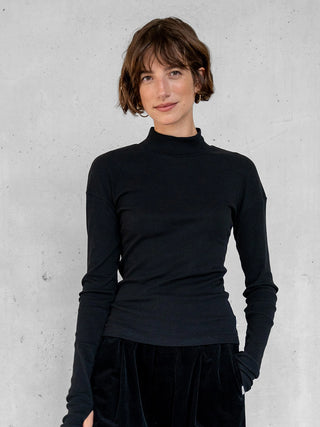 Junge Frau in Penn&Ink Longsleeve schwarz: eng anliegend mit Stehkragen und langen Ärmeln, aus weichem, elastischem Material, vor einer hellgrauen Betonwand stehend, lächelnd.