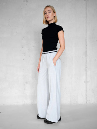 Junge Frau trägt das Penn&Ink Top black, ein figurbetontes Kurzarm-Top mit Stehkragen, kombiniert mit weitem, hellgrauem Hose. Sie posiert lässig vor einer minimalistischen Betonwand.