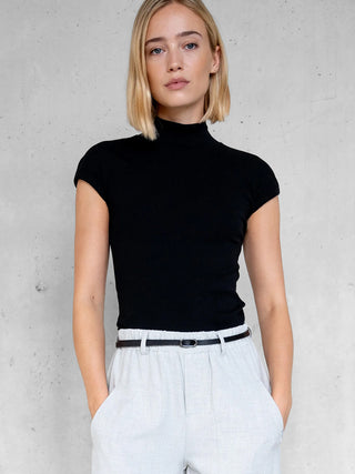 Penn&Ink Top black: Junge Frau trägt ein schwarzes, figurbetontes Kurzarm-Turtleneck-Top, kombiniert mit hellgrauen, hoch taillierten Hosen. Der minimalistische Look betont die elegante Schlichtheit des Oberteils.