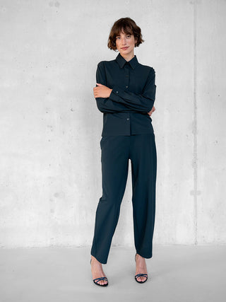Die Penn&Ink Bluse antra wird an einer schlanken Person gezeigt, die gegen eine graue Wand steht. Sie trägt die dunkelknöpfbare Bluse mit klassischem Hemdkragen, kombiniert mit passenden Hosen und offenen Absätzen.