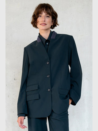 Penn&Ink Blazer antra: Junge Frau im dunkel geschnittenen Blazer mit kontrastierendem Kragen, vier Knöpfen und Pattentaschen, zeigt elegante, moderne Silhouette vor einer strukturierten Wand. Trägt goldene Creolen.