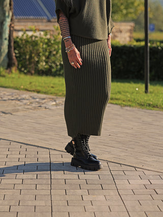 Person trägt den OFF#DLY Rock Linda green, einen gerippten Strickrock in Midi-Länge, kombiniert mit schwarzen Schnürstiefeln. Armbänder und Ringe akzentuieren den Look im parkähnlichen Umfeld.