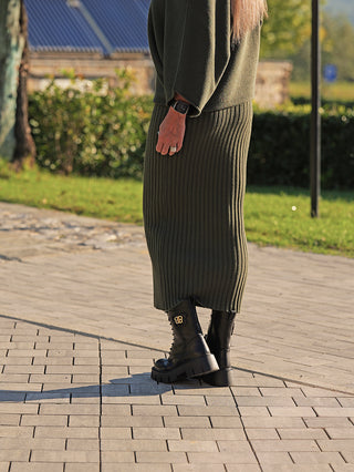 Person trägt den OFF#DLY Rock Linda green, einen olivgrünen, gerippten Midi-Rock, kombiniert mit passendem Pullover. Dazu schwarze, klobige Stiefeletten mit BB-Logo und einem Fitness-Tracker.