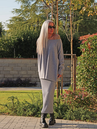 Person trägt OFF#DLY Rock Linda grey, einen gerippten, grauen Midi-Strickrock, kombiniert mit einem grauen Pullover und schwarzen Combat-Boots, im Garten mit grünen Büschen und blühenden Sträuchern.